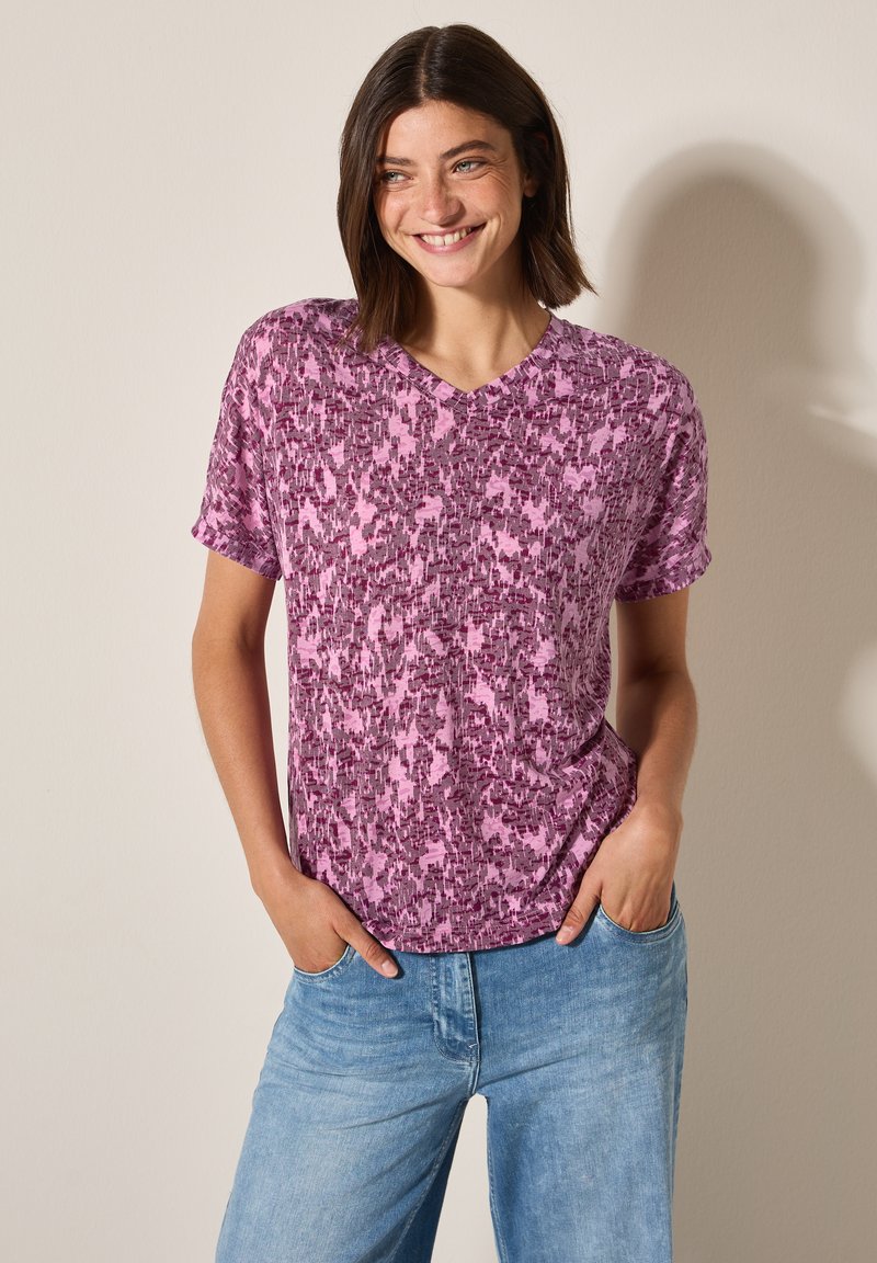 T-shirt à imprimé graphique violet et rose avec des manches courtes et un col en V. Tissu doux, coupe décontractée, associé à un jean bleu clair.