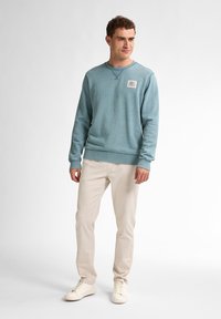 Felpa azzurra con maniche lunghe e scollatura rotonda, caratterizzata da una piccola toppa sul petto. Abbinata a pantaloni beige e sneakers bianche.