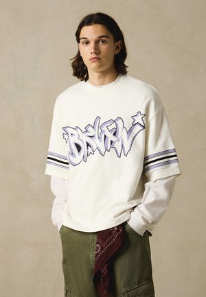 Mladá osoba s dlouhými vlasy nosící bílou oversized košili s graffiti textem "BRKLN" a olivově zelené cargo kalhoty s visícím červeným šátkem.