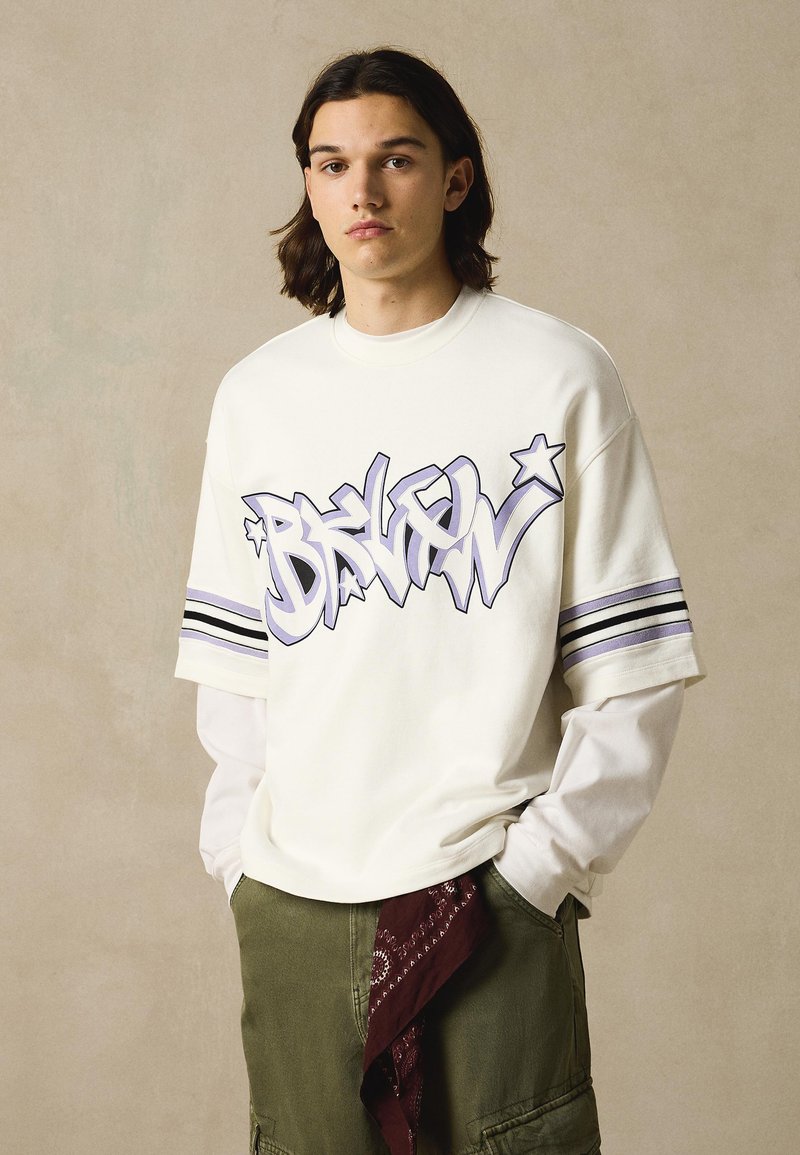 Junge Person mit langen Haaren, die ein übergroßes weißes Hemd mit Graffiti-Schriftzug "BRKLN" und olivgrüne Cargohosen mit einem hängenden roten Bandana trägt.