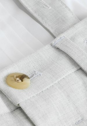 Lysegråt stof med en tekstureret vævning, med en stor, rund, lys knap. Nærbillede af stropfests detalje.