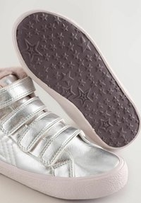Silberne metallische Sneakers mit drei Klettverschlüssen, strukturiertem weißen Gummisohle und lila Außensohle mit Sternmuster. Weiche Innenauskleidung sichtbar.