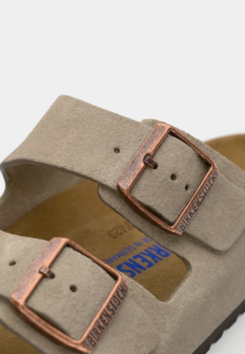 Light grey suede sandals with two adjustable straps, featuring copper-colored metal buckles, and a textured footbed.