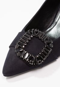Gros plan d'une chaussure en daim noir à bout pointu avec une grande pierre noire carrée en décoration à l'avant.