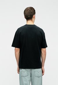 Μαύρο βαμβακερό μπλουζάκι με κοντά μανίκια, άνετη εφαρμογή και κυκλή λαιμόκοψη, φορεμένο με ανοιχτό μπλε χαλαρούς τζιν παντελόνες.