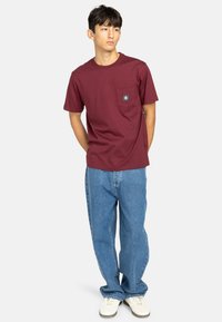 Camiseta de manga corta burdeos con un bolsillo en el pecho, combinada con unos vaqueros sueltos de un azul claro y zapatillas blancas. Diseño minimalista.
