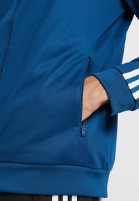 Modrá zipová bunda s hladkou texturou, se bočními zipovými kapsami a bílými pruhy na rukávech.