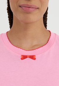 Rózsaszín póló, fodros vörös nyakkivágással és egy kis vörös masnival a mellrészen. Sima pamut anyag. Közeli nézet.
