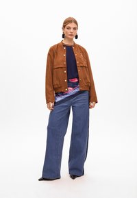 Brązowa zamszowa kurtka bombowiec z zatrzaskami, granatowa bluzka i szerokie niebieskie dżinsy. Czarne spiczaste botki do kostek dodają kontrastu. Gładkie tekstury.