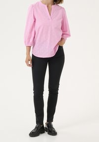 Blouse rose clair avec un décolleté en trou de serrure, des manches bouffantes et des motifs texturés, associée à un jean noir slim et des chaussures noires.