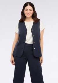 Marineblauwe mouwloze vest met vijf zilveren knopen, gecombineerd met een witte korte mouwen shirt en bijpassende marinebroeken. Glad stoffen textuur.