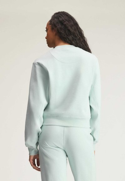 Kobieta o długich kręconych włosach, ubrana w jasnoniebieską bluzę i dopasowane spodnie, stojąca tyłem na jednolitym tle.