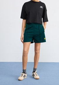 Svart kortärmad t-shirt med logga, tillsammans med gröna träningsshorts med sidfickor och gula detaljer; beige sneakers med svarta ränder.