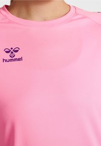 Różowa koszulka sportowa z gładkiego materiału, z okrągłym dekoltem i fioletowym logotypem "hummel" na lewej piersi.