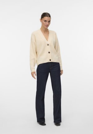 Cardigan a coste crema con scollo a V, decorato con tre grandi bottoni marroni. Abbinato a jeans a palazzo blu scuro. Design semplice e testurizzato.