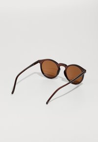  Runde solbriller med mørkebrune rammer, brune tonede linser og tynne stenger. Designet er minimalistisk med en glatt finish.