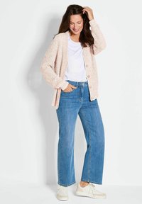 Lichtroze gebreide cardigan met knopen, gedragen over een wit T-shirt en gecombineerd met blauwe wijde jeans en witte sneakers. Gladde stoftextuur.