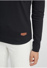 Camiseta negra de manga larga en material suave con dobladillo redondeado, que presenta una pequeña etiqueta de cuero marrón con "oxmo" cerca de la parte inferior.