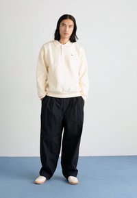 Κρέμα φούτερ με γιακά και ελαστική μέση, σε συνδυασμό με χαλαρούς μαύρους παντελόνες. Υποδήματα είναι αθλητικά παπούτσια σε ανοιχτό χρώμα. Μινιμαλιστικός σχεδιασμός.