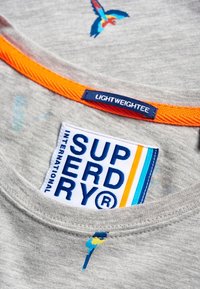 Lehká šedá bavlněná tričko s oranžovým límečkem, vyšitým ptáčkem a bílou etiketou s nápisem "SUPERDRY" v modré barvě a barevnými pruhy.