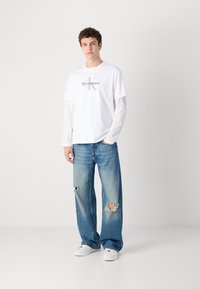 Bijela grafička majica s "Calvin Klein" logom, složena preko bijele košulje dugih rukava, u kombinaciji s svijetloplavim iznošenim širokim trapericama i bijelim tenisicama.
