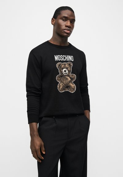 Άντρας που φοράει μαύρο πουλόβερ Moschino με εικόνα αρκουδάκι και μαύρο παντελόνι, στέκεται με το ένα χέρι στην τσέπη μπροστά σε απλό φόντο.