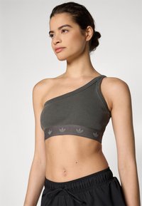 Asymmetrisk mörkgrå ribbad sport-bh med ett brett elastiskt band som pryds av ett upprepat logomönster. Mjuk textur med en åtsittande design.