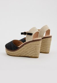 Sandalias de cuña en negro y beige con una correa ajustable en el tobillo, parte superior de tela texturizada y detalles de plataforma de cuerda trenzada.