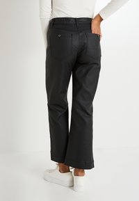 Persona vestida con pantalones de pierna ancha negros con detalles de botones dorados en los bolsillos traseros y zapatillas blancas, de pie contra un fondo blanco.