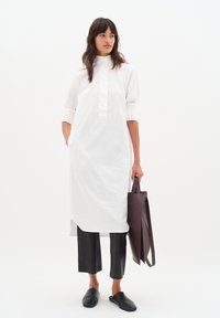 Weißes Langarmhemdkleid mit hohem Kragen, kombiniert mit schwarzen verkürzten Hosen und schwarzen Slippern, während sie eine dunkelbraune Tote-Bag hält.