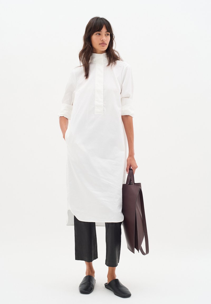 Weißes Langarmhemdkleid mit hohem Kragen, kombiniert mit schwarzen verkürzten Hosen und schwarzen Slippern, während sie eine dunkelbraune Tote-Bag hält.