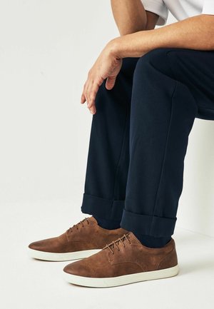 Personne assise, les mains reposant sur les genoux, portant un pantalon bleu marine et des chaussures décontractées marron à lacets avec des semelles blanches.