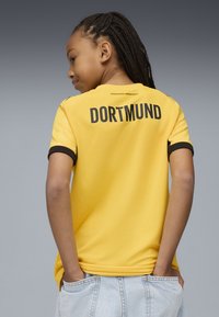 Otrok z zapletom v laseh, oblečen v rumeno nogometno majico Borussia Dortmund, z rokami v žepih, obrnjeno stran od enobarvne sive podlage.