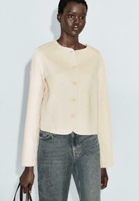 Donna con capelli corti che indossa una giacca beige con bottoni e jeans blu sbiaditi, mentre tiene una borsa marrone su uno sfondo grigio chiaro.