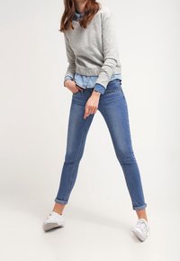 Felpa grigia chiara indossata sopra una camicia di denim azzurro, abbinata a jeans skinny blu e sneakers bianche. Esibizione di un outfit casual.