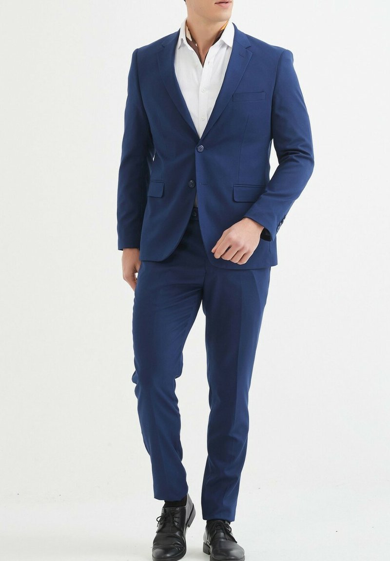 Costume bleu marine en tissu lisse, comprenant un blazer à simple bouton avec deux boutons, et un pantalon slim assorti. Chaussures noires à lacets.
