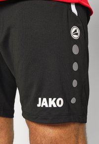 Schwarze Sportshorts aus leichtem Material, mit einem weißen Logo "JAKO" und kreisförmigen Punktakzenten an der Seite.