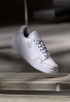 White Adidas sneaker with laces and trefoil logo, suspended above a textured stone bench in soft indoor lighting.