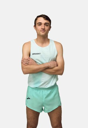 Jonge man staat met gekruiste armen, draagt een lichtblauw Brooks hardloopsingletje en bijpassende korte broek tegen een effen achtergrond.