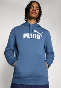 Blaues Kapuzensweatshirt mit großem weißen "PUMA"-Logo und einem kleineren Logo darüber. Mit einer Fronttasche und strukturierten Bündchen. Aus weichem Material.