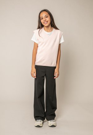 Fille souriante aux longs cheveux bruns, portant un pull sans manches rose clair par-dessus un t-shirt blanc, un pantalon large noir et des baskets blanches.