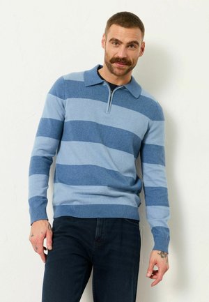 Mann mit kurzem Haar und Schnurrbart, der einen blau gestreiften Pullover und dunkle Hosen trägt, steht vor einer hellen Wand, die Hände entspannt an den Seiten.