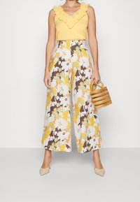 Gele gerimpelde tanktop gecombineerd met een broek met wijde pijpen met een bloemenpatroon in geel, bruin en wit. Accessoire: kleine geweven handtas.