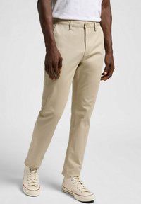 Chinos caqui con corte recto, hechas de una mezcla de algodón. Cuenta con un cierre de botón, trabillas para el cinturón y piernas ajustadas, combinados con zapatillas beige.