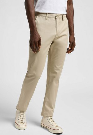 Khaki Chinos mit geradem Schnitt, aus einem Baumwollmischgewebe gefertigt. Sie verfügen über einen Knopfverschluss, Gürtelschlaufen und schmal zulaufende Beine, die mit beigen Sneakers kombiniert sind.