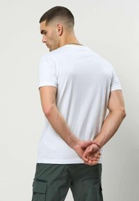 Homme en chemise blanche à manches courtes et pantalon cargo foncé, debout de dos à la caméra, mains jointes derrière lui, tête tournée vers la gauche.