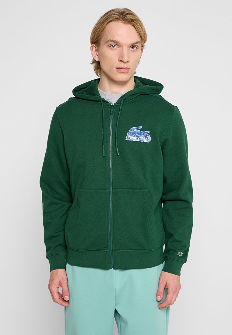 Haut à zip vert en tissu mélangé coton. Comprend une poche avant et un logo bleu brodé sur la poitrine gauche. Capuche avec cordon de serrage.