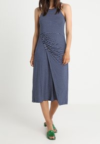 Granatowa sukienka w paski z dekoltem na szyi i marszczonym detalem w talii, o długości do kolan i bocznym rozcięciu. Zestawiona z zielonymi klapkami.