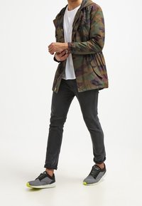 Osoba kráčející v maskáčové bundě, bílé tričko, tmavé slim džíny a šedé sneakers s neónově zelenými podrážkami.