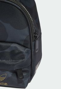 Black camo backpack with textured fabric, zip closure, and black hardware. Features gold logo on the front and a side pocket accent.